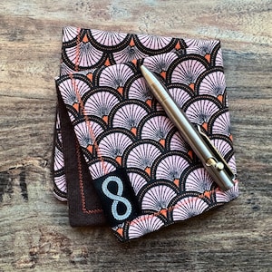 May include: Two fabric squares with a pink and black geometric pattern. The fabric squares are folded and a silver pen is lying on top of them. The fabric squares have a brown fabric border with an infinity symbol in the centre.