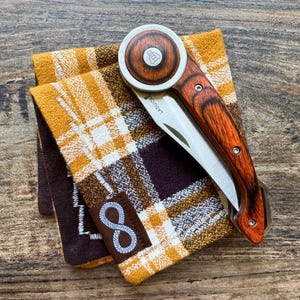 Könnte beinhalten: Ein braunes und silbernes Taschenmesser mit einem Holzgriff und einer silbernen Klinge. Das Messer liegt auf einem braunen und gelben karierten Stoff.
