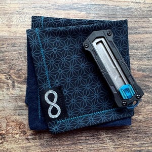 Könnte beinhalten: Ein dunkelblaues, gemustertes Stofftaschentuch mit einem schwarzen und blauen Universalmesser und einem weißen Patch mit der Nummer "8" darauf.