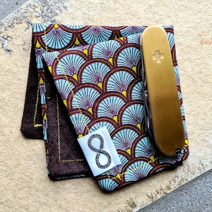 Könnte beinhalten: Ein braunes und blaues gemustertes Stofftaschentuch mit einem goldenen Taschenmesser und einem weißen Etikett mit der Nummer "8" darauf.