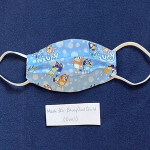 May include: A blue and white fabric face mask with a pattern of cartoon dogs and bubbles. The mask has white elastic ear loops and the text "Bluey" is visible on the mask. The mask is labeled "Mask 301 - Bluey Oval Child (Oval)".