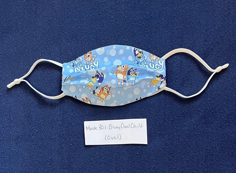 May include: A blue and white fabric face mask with a pattern of cartoon dogs and bubbles. The mask has white elastic ear loops and the text "Bluey" is visible on the mask. The mask is labeled "Mask 301 - Bluey Oval Child (Oval)".
