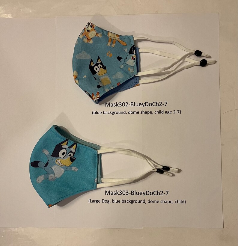 May include: Two blue fabric face masks with a cartoon dog print. The top mask has a smaller dog and the bottom mask has a larger dog. Both masks have white elastic straps and black toggles. The text on the image reads "Mask302-BlueyDoCh2-7 (blue background, dome shape, child age 2-7)" and "Mask303-BlueyDoCh2-7 (Large Dog, blue background, dome shape, child)"