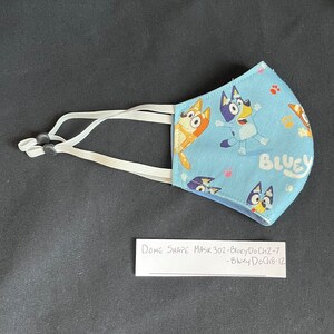 May include: Blue and white fabric face mask with a cartoon dog design. The mask has elastic ear loops and the text "Bluey" is printed on the mask. The text "Dome Shape Mask 30z - Bluey DoCh2-7 - Bluey DoCh8-12" is written on a white piece of paper.