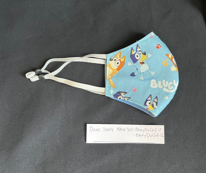 May include: Blue and white fabric face mask with a cartoon dog design. The mask has elastic ear loops and the text "Bluey" is printed on the mask. The text "Dome Shape Mask 30z - Bluey DoCh2-7 - Bluey DoCh8-12" is written on a white piece of paper.