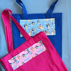 May include: Two tote bags, one blue and one pink, with a patterned fabric insert featuring cartoon dogs. The pink bag has the text "Bluey" on the insert.