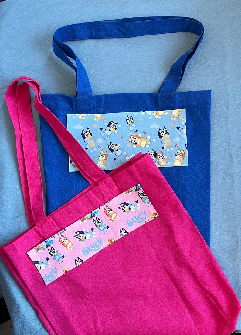 May include: Two tote bags, one blue and one pink, with a patterned fabric insert featuring cartoon dogs. The pink bag has the text "Bluey" on the insert.