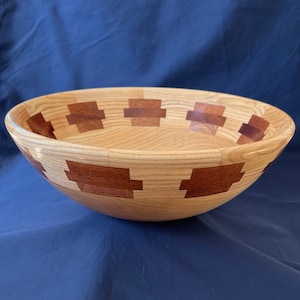 K&ouml;nnte beinhalten: Eine gro&szlig;e Holzschale mit einer nat&uuml;rlichen Holzoptik und einer dekorativen Einlage aus r&ouml;tlich-braunem Holz. Die Schale hat eine abgerundete Form und einen breiten Rand.
