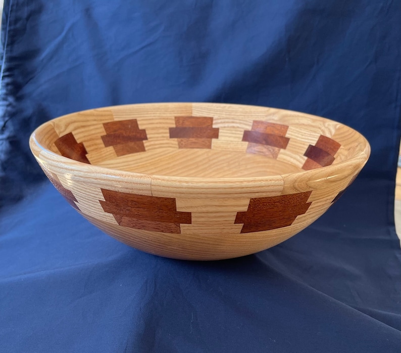 K&ouml;nnte beinhalten: Eine gro&szlig;e Holzschale mit einer nat&uuml;rlichen Holzoptik und einer dekorativen Einlage aus r&ouml;tlich-braunem Holz. Die Schale hat eine abgerundete Form und einen breiten Rand.