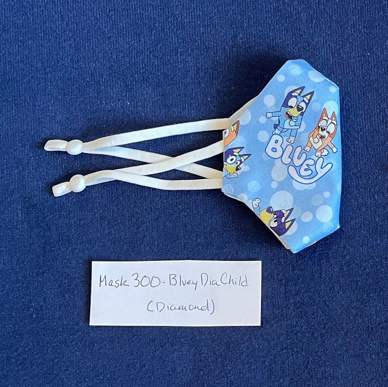 May include: Bluey face mask for children with a blue background and white polka dots. The mask has a white elastic band and features the characters Bluey and Bingo. The text "Bluey" is printed on the mask. The mask is labeled "Mask 300 - Bluey Dia Child (Diamond)".