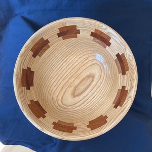 K&ouml;nnte beinhalten: Eine Holzsch&uuml;ssel mit nat&uuml;rlicher Holzoptik und einem dekorativen Rand aus eingelegtem Holz in rotbrauner Farbe.