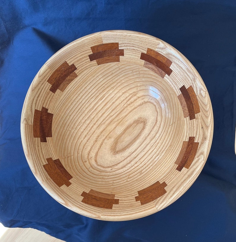 K&ouml;nnte beinhalten: Eine Holzsch&uuml;ssel mit nat&uuml;rlicher Holzoptik und einem dekorativen Rand aus eingelegtem Holz in rotbrauner Farbe.