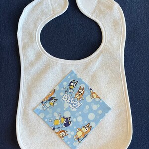 May include: White terry cloth baby bib with a blue and orange cartoon character print fabric square on top. The fabric square has the word "Bluey" printed on it.