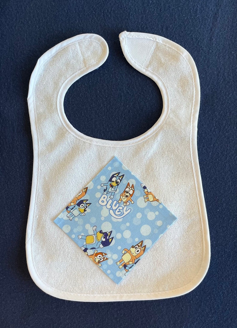 May include: White terry cloth baby bib with a blue and orange cartoon character print fabric square on top. The fabric square has the word "Bluey" printed on it.