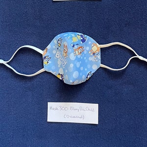 May include: Blue and white face mask with a cartoon dog print. The mask has white elastic straps and a label that reads "Mask 300 - Bluey Dia Child (Discontinued)".