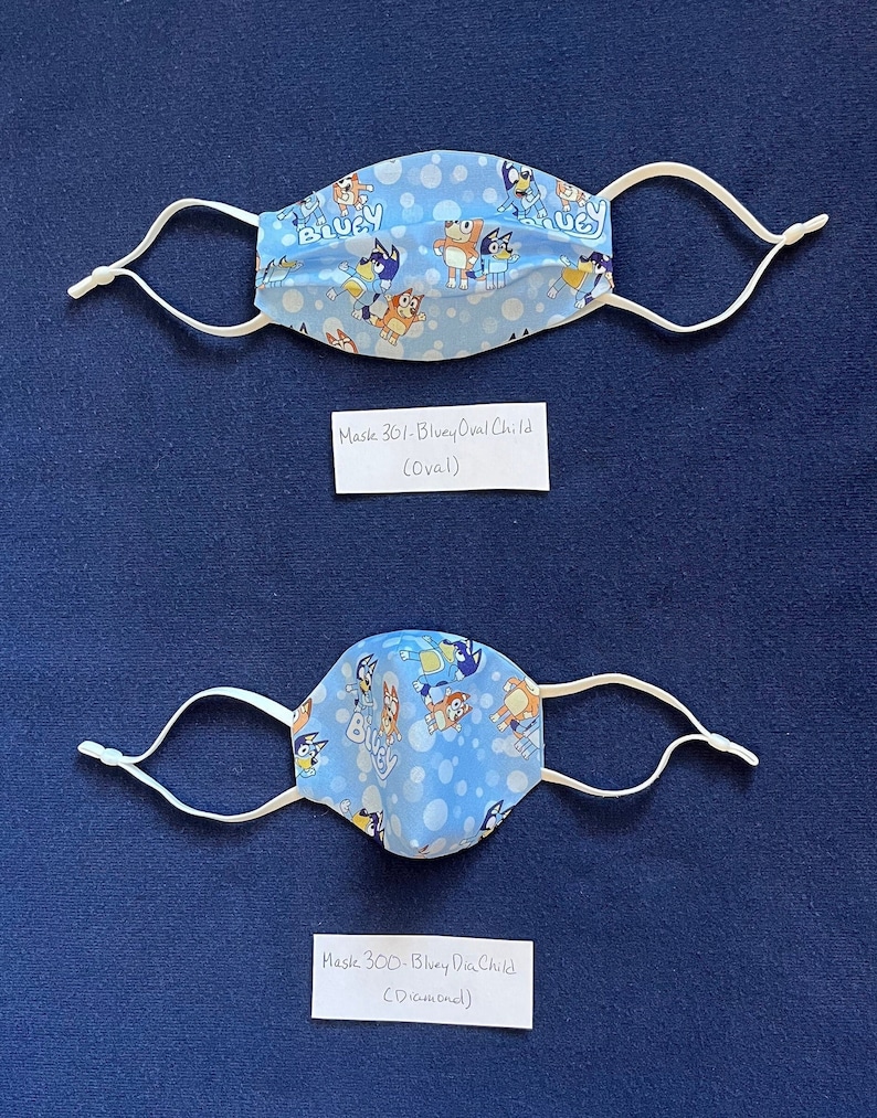 May include: Two blue and white fabric face masks with elastic ear loops. The masks feature a cartoon dog character with the word "Bluey" on the fabric. One mask is oval shaped and the other is diamond shaped. The text "Mask 361-Bluey Oval Child (Oval)" and "Mask 300-Bluey Dia Child (Diamond)" is written on white cards below each mask.