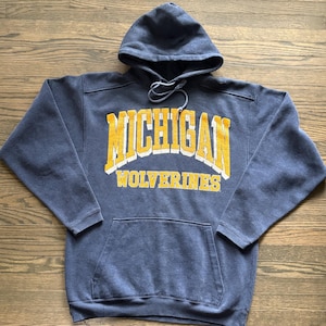 Peut inclure: Un sweat-shirt à capuche bleu marine arborant le mot MICHIGAN en grandes lettres jaunes avec des contours blancs, et le mot WOLVERINES en plus petites lettres jaunes en dessous. Le sweat possède une poche kangourou et des cordons.