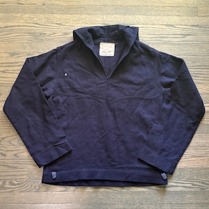 Könnte beinhalten: Ein dunkelblaues Langarm-Pullover-Shirt mit einem breiten Matrosenkragen, einem V-Ausschnitt und einer horizontalen Naht über der Brust. Ein rechteckiges weißes Stoffetikett ist am inneren Halsausschnitt angenäht und ein kleines Loch ist sichtbar.