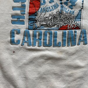 Może przedstawiać: Biała bluza z napisem "NORTH CAROLINA" w jasnoniebieskich literach blokowych. Projekt zawiera koszyk&oacute;wkę, numer "84" i słowo "CAROLINA" w kolorze jasnoniebieskim. Bluza ma okrągły dekolt.
