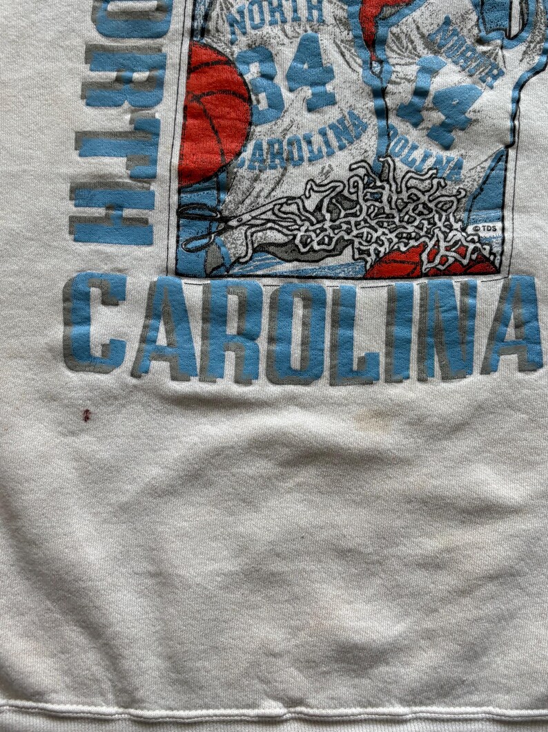 Może przedstawiać: Biała bluza z napisem "NORTH CAROLINA" w jasnoniebieskich literach blokowych. Projekt zawiera koszyk&oacute;wkę, numer "84" i słowo "CAROLINA" w kolorze jasnoniebieskim. Bluza ma okrągły dekolt.