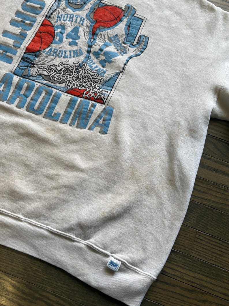 Może przedstawiać: Kremowa bluza z nadrukiem koszulki do koszyk&oacute;wki z napisem "North Carolina" i numerem "34" w kolorze niebieskim. Projekt zawiera koszyk&oacute;wkę i kontur mapy. Bluza ma prążkowany d&oacute;ł i mankiety.