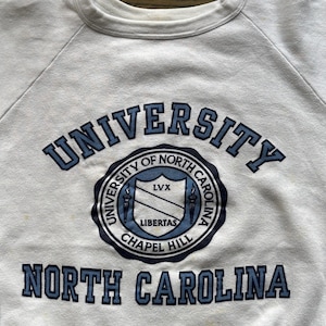 Pu&ograve; includere: Felpa bianca girocollo con scritte blu e un emblema circolare. Il testo recita "UNIVERSITY OF NORTH CAROLINA" e "NORTH CAROLINA". L'emblema include "LIBERTAS" e "CHAPEL HILL".