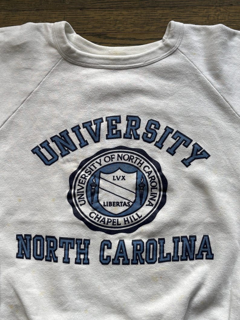 Pu&ograve; includere: Felpa bianca girocollo con scritte blu e un emblema circolare. Il testo recita "UNIVERSITY OF NORTH CAROLINA" e "NORTH CAROLINA". L'emblema include "LIBERTAS" e "CHAPEL HILL".
