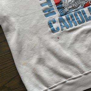 Może przedstawiać: Jasnoszara bluza z napisem "NORTH CAROLINA" w jasnoniebieskich literach blokowych. Grafika zawiera koszyk&oacute;wkę i inne obrazy. Bluza ma okrągły dekolt oraz prążkowane mankiety i d&oacute;ł.