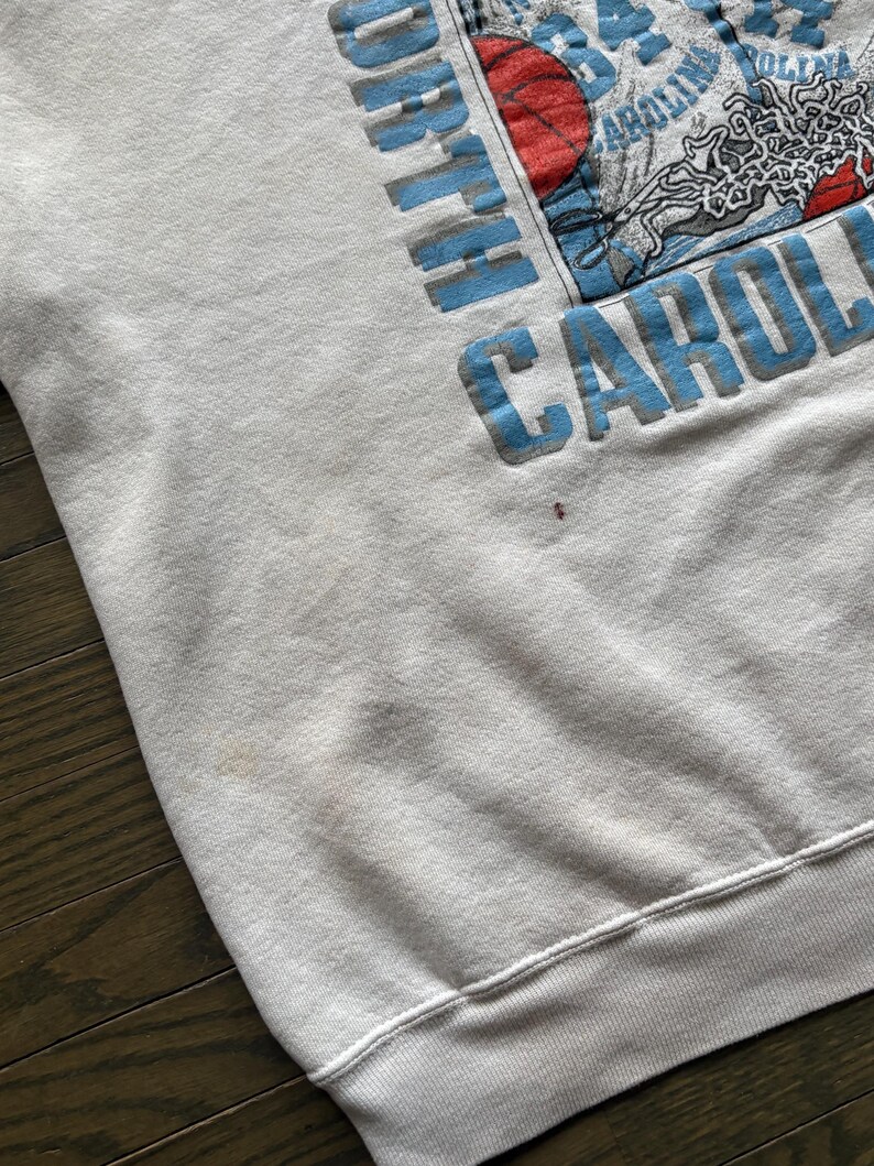 Może przedstawiać: Jasnoszara bluza z napisem "NORTH CAROLINA" w jasnoniebieskich literach blokowych. Grafika zawiera koszyk&oacute;wkę i inne obrazy. Bluza ma okrągły dekolt oraz prążkowane mankiety i d&oacute;ł.