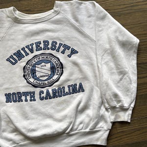 Pu&ograve; includere: Felpa bianca girocollo con le parole "UNIVERSITY OF NORTH CAROLINA CHAPEL HILL" in lettere blu. La felpa ha un emblema circolare con le parole "LIBERTAS" e "LVX" all'interno. La felpa &egrave; realizzata in materiale morbido.