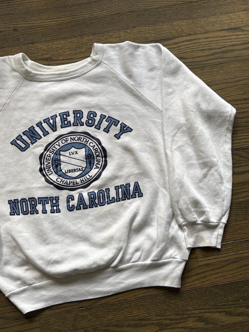 Pu&ograve; includere: Felpa bianca girocollo con le parole "UNIVERSITY OF NORTH CAROLINA CHAPEL HILL" in lettere blu. La felpa ha un emblema circolare con le parole "LIBERTAS" e "LVX" all'interno. La felpa &egrave; realizzata in materiale morbido.