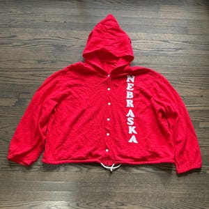 Puede incluir: Una chaqueta con capucha de color rojo brillante con cierre de botones frontal y mangas largas. La palabra NEBRASKA está impresa verticalmente en letras mayúsculas blancas a lo largo del lado derecho de la tapeta. Un cordón blanco es visible en el dobladillo.