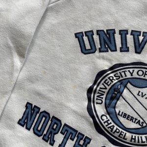 Pu&ograve; includere: Felpa bianca con la scritta blu "UNIVERSITY OF NORTH CAROLINA CHAPEL HILL". &Egrave; visibile anche il sigillo dell'universit&agrave;, con uno scudo e la parola "LIBERTAS".