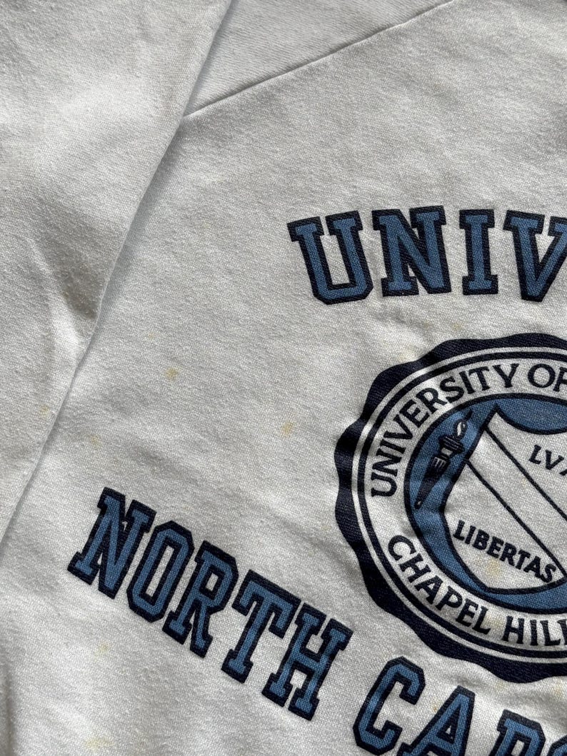 Pu&ograve; includere: Felpa bianca con la scritta blu "UNIVERSITY OF NORTH CAROLINA CHAPEL HILL". &Egrave; visibile anche il sigillo dell'universit&agrave;, con uno scudo e la parola "LIBERTAS".