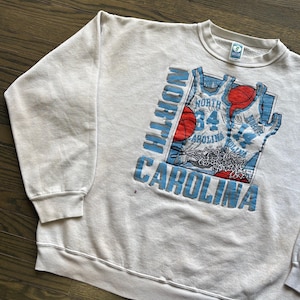 Może przedstawiać: Kremowa bluza w stylu vintage z nadrukiem koszykarskim. Grafika zawiera napisy "NORTH CAROLINA" w jasnoniebieskim kolorze, wz&oacute;r koszulki do koszyk&oacute;wki oraz czerwone i jasnoniebieskie piłki do koszyk&oacute;wki. Bluza ma okrągły dekolt i długie rękawy.