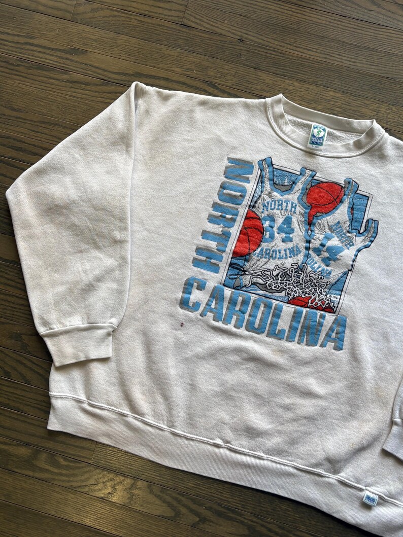 Może przedstawiać: Kremowa bluza w stylu vintage z nadrukiem koszykarskim. Grafika zawiera napisy "NORTH CAROLINA" w jasnoniebieskim kolorze, wz&oacute;r koszulki do koszyk&oacute;wki oraz czerwone i jasnoniebieskie piłki do koszyk&oacute;wki. Bluza ma okrągły dekolt i długie rękawy.