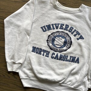Pu&ograve; includere: Felpa bianca girocollo con la scritta "UNIVERSITY OF NORTH CAROLINA" in blu navy. La felpa presenta un emblema circolare con le parole "UNIVERSITY OF NORTH CAROLINA CHAPEL HILL" e la parola "LIBERTAS".