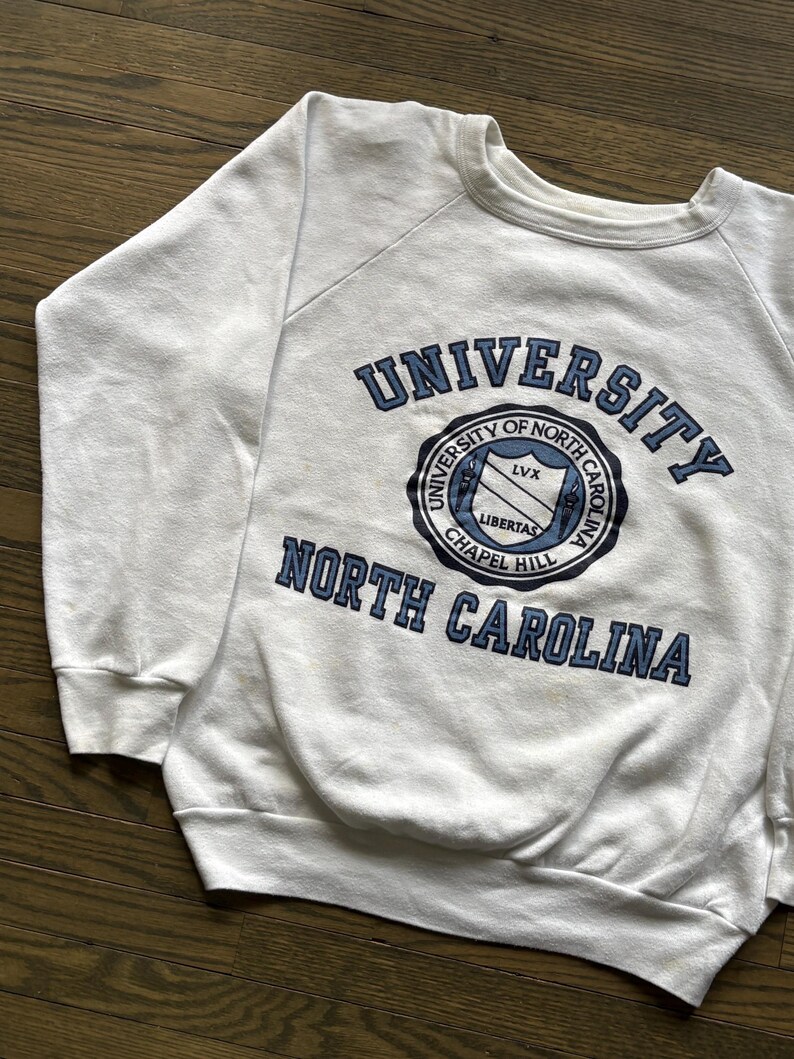 Pu&ograve; includere: Felpa bianca girocollo con la scritta "UNIVERSITY OF NORTH CAROLINA" in blu navy. La felpa presenta un emblema circolare con le parole "UNIVERSITY OF NORTH CAROLINA CHAPEL HILL" e la parola "LIBERTAS".