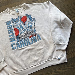 Może przedstawiać: Kremowa bluza z nadrukiem koszykarskim. Grafika zawiera słowa "North Carolina" w kolorze niebieskim, piłki do koszyk&oacute;wki i wzory koszulek. Bluza ma okrągły dekolt i długie rękawy.
