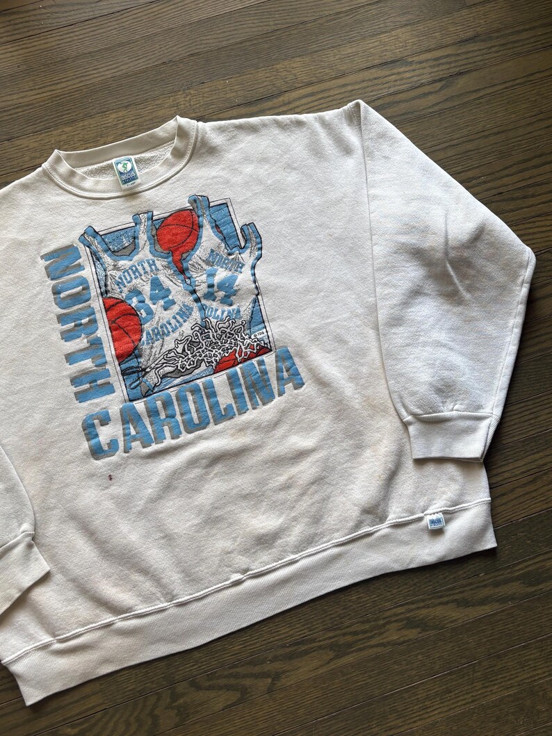 Może przedstawiać: Kremowa bluza z nadrukiem koszykarskim. Grafika zawiera słowa "North Carolina" w kolorze niebieskim, piłki do koszyk&oacute;wki i wzory koszulek. Bluza ma okrągły dekolt i długie rękawy.