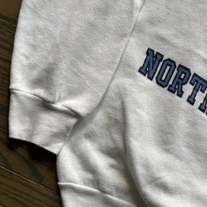 Pu&ograve; includere: Felpa bianca con la scritta "NORTH" in lettere maiuscole blu navy. La felpa ha girocollo e maniche lunghe. Il tessuto appare leggermente usurato.