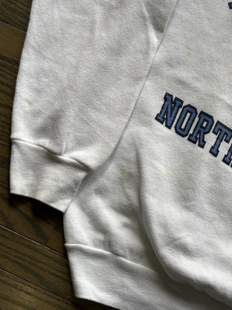 Pu&ograve; includere: Felpa bianca con la scritta "NORTH" in lettere maiuscole blu navy. La felpa ha girocollo e maniche lunghe. Il tessuto appare leggermente usurato.