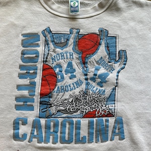 Może przedstawiać: Kremowa bluza z okrągłym dekoltem z grafiką o tematyce koszyk&oacute;wki. Projekt zawiera koszulki do koszyk&oacute;wki i piłki, z napisem "NORTH CAROLINA" w kolorze niebieskim. Odzież w stylu vintage.