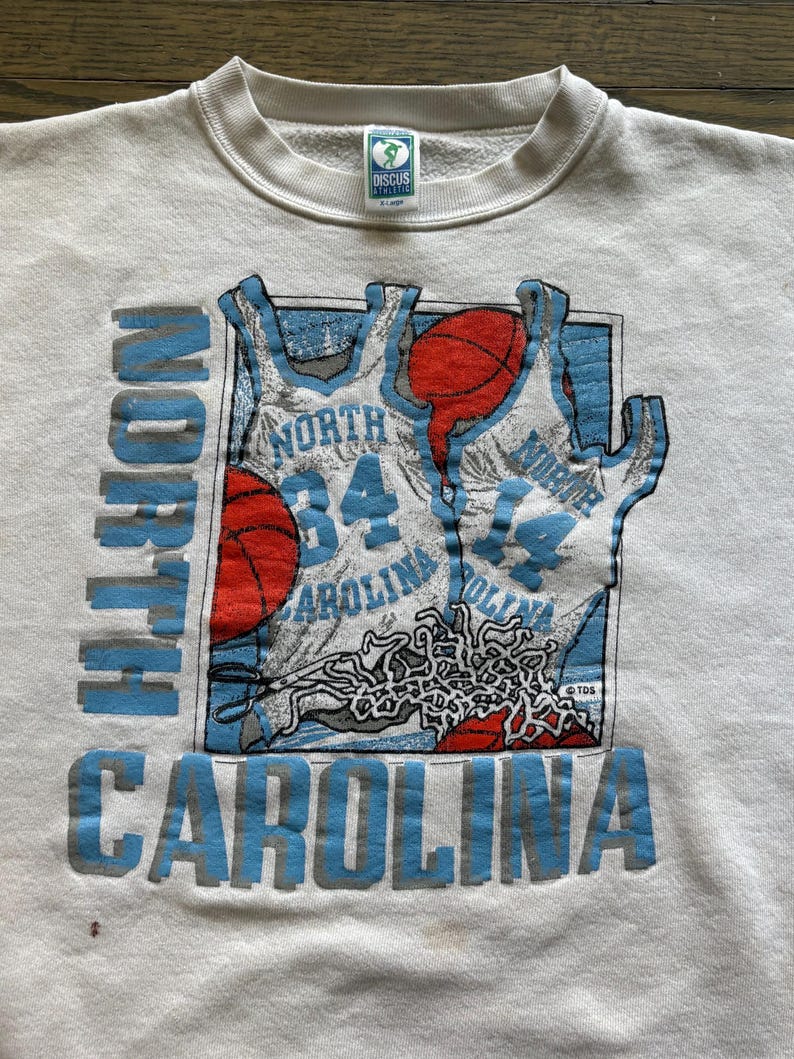 Może przedstawiać: Kremowa bluza z okrągłym dekoltem z grafiką o tematyce koszyk&oacute;wki. Projekt zawiera koszulki do koszyk&oacute;wki i piłki, z napisem "NORTH CAROLINA" w kolorze niebieskim. Odzież w stylu vintage.