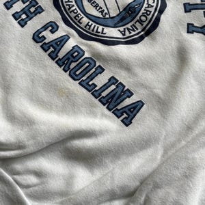 Pu&ograve; includere: Felpa bianca con le parole "NORTH CAROLINA" in lettere blu. Un emblema circolare con "UNIVERSITY OF NORTH CAROLINA CHAPEL HILL" circonda uno scudo con la parola "LIBERTAS".
