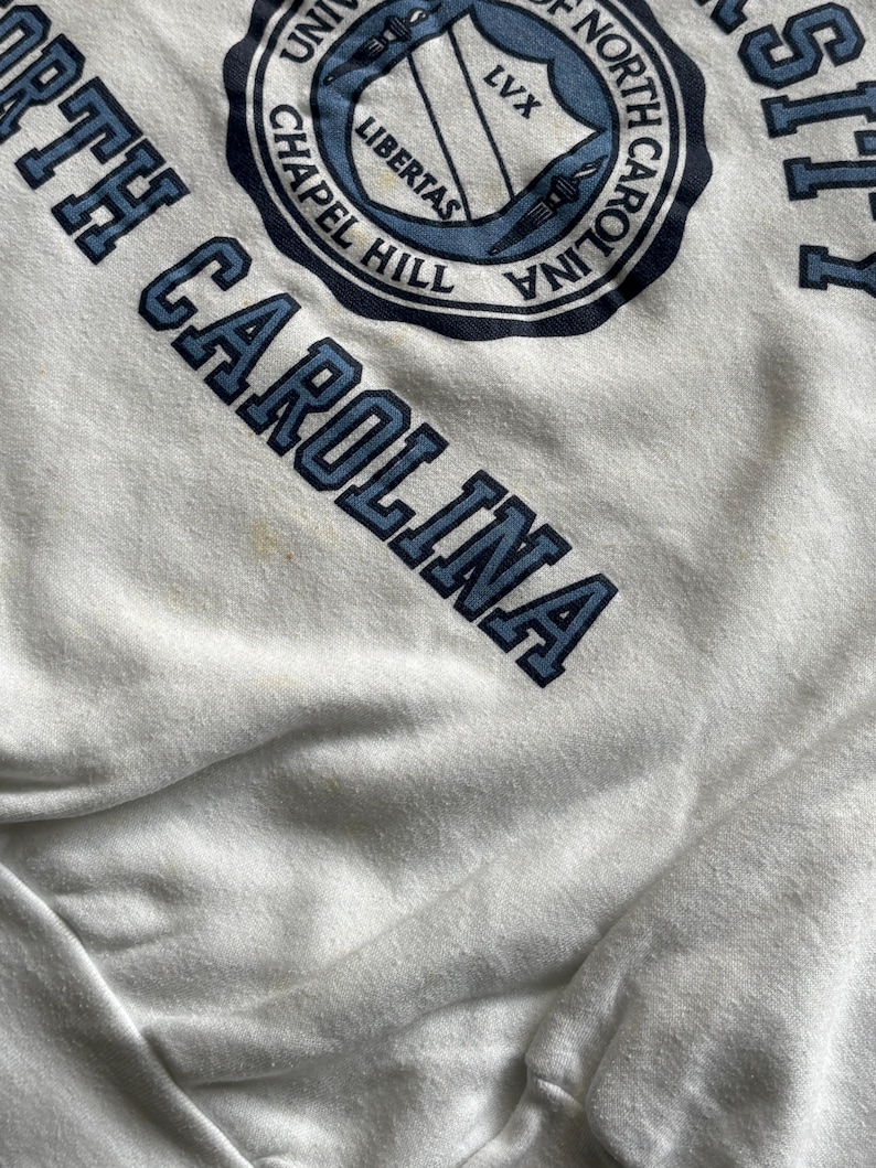 Pu&ograve; includere: Felpa bianca con le parole "NORTH CAROLINA" in lettere blu. Un emblema circolare con "UNIVERSITY OF NORTH CAROLINA CHAPEL HILL" circonda uno scudo con la parola "LIBERTAS".