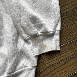 Pu&ograve; includere: Primo piano di una manica di felpa bianca con polsino a coste. Il tessuto mostra segni di usura e scolorimento, con macchie visibili sul polsino. Lo sfondo &egrave; una superficie di legno.