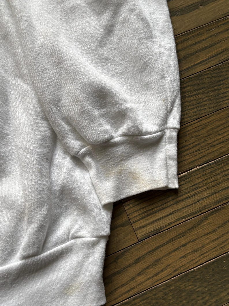 Pu&ograve; includere: Primo piano di una manica di felpa bianca con polsino a coste. Il tessuto mostra segni di usura e scolorimento, con macchie visibili sul polsino. Lo sfondo &egrave; una superficie di legno.