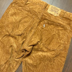 May include: Close-up of a pair of tan corduroy pants. The pants feature two back pockets and a Levi Strauss & Co. label. The corduroy fabric has a ribbed texture.