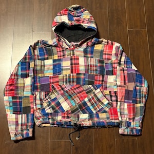 May include: A colourful patchwork hoodie with a drawstring waist and a front pocket. The hood is lined with black fabric. The patchwork design incorporates red, blue, and purple squares. The hoodie is a casual garment.